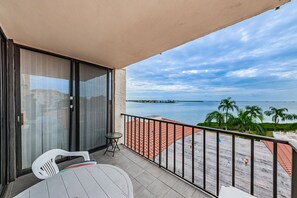 Un balcón con puerta de vidrio, una mesita y dos sillas. Se ve una playa con palmeras y una extensión de agua.