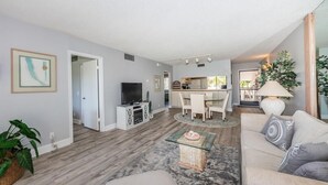 A modern living room with a sofa, coffee table, and TV. There is a dining area with chairs and a table. The room has wooden flooring and a large window. - 2BR condo with golf course views, pool, hot tub, and balcony. (St. Petersburg)