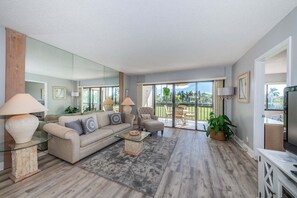Una sala moderna con sofá, sillones y una mesita de centro. Hay un balcón con mobiliario exterior y vistas a zonas verdes.