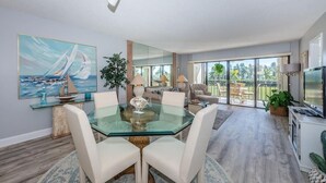 A spacious living room with a glass dining table, comfortable seating, a large mirror, and a view of the outdoors. - 2BR condo with golf course views, pool, hot tub, and balcony. (St. Petersburg)