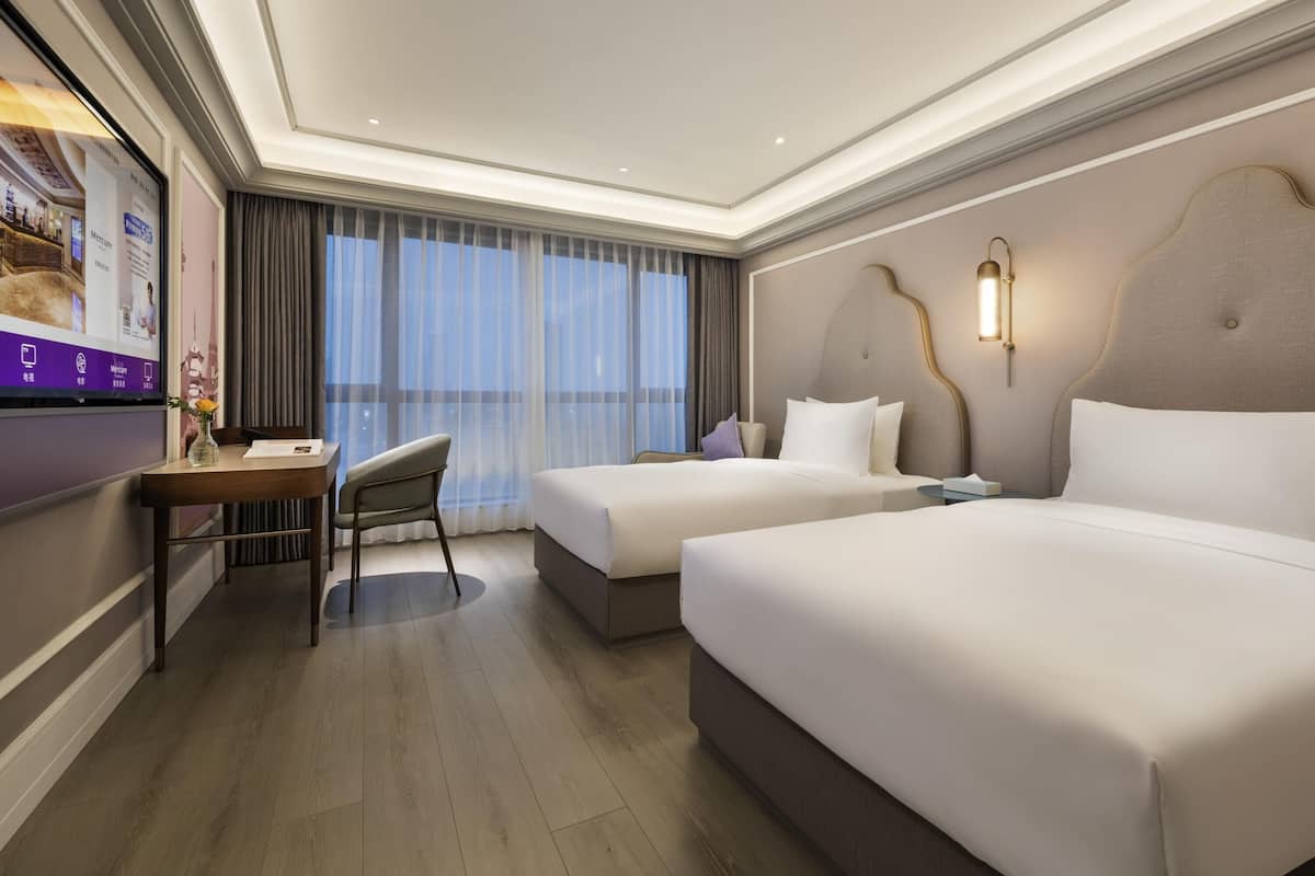 A hotel room with two beds, a desk with a chair, a TV, and a large window with curtains.