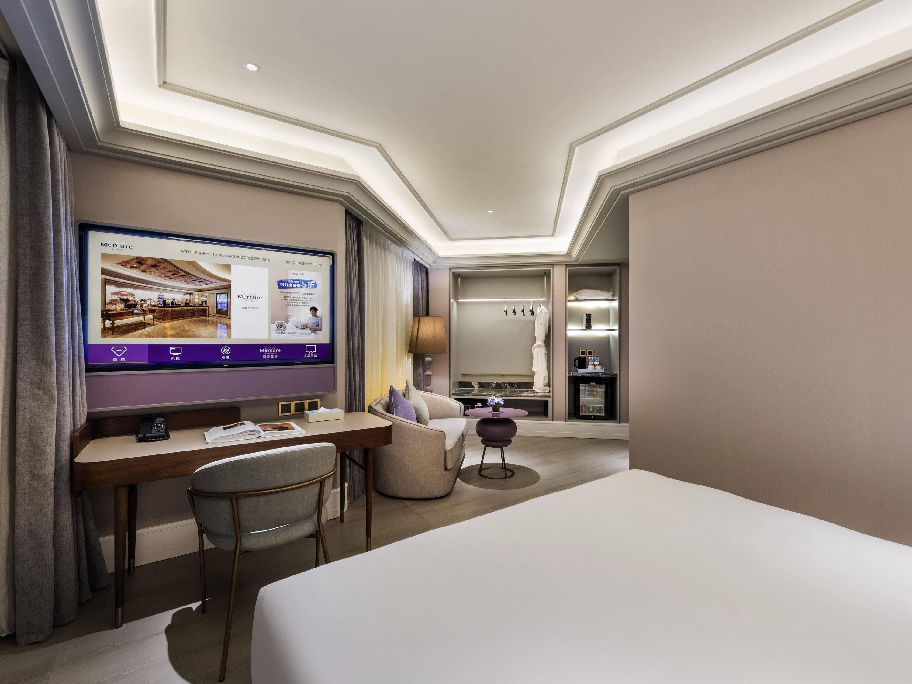 A hotel room with a large flat-screen TV, a desk with a chair, a bed, a sofa, and a small table with a lamp.