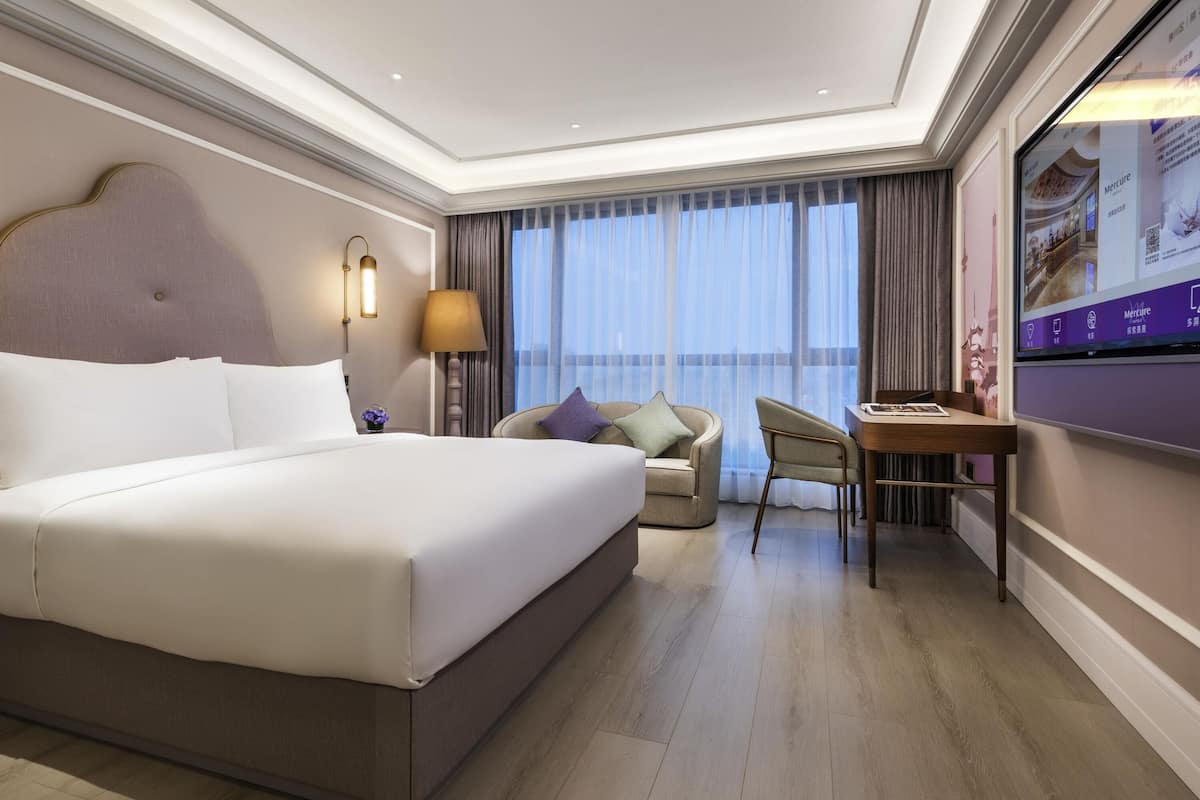 A hotel room with a large bed, a desk, a chair, a TV, and a view of the outdoors.