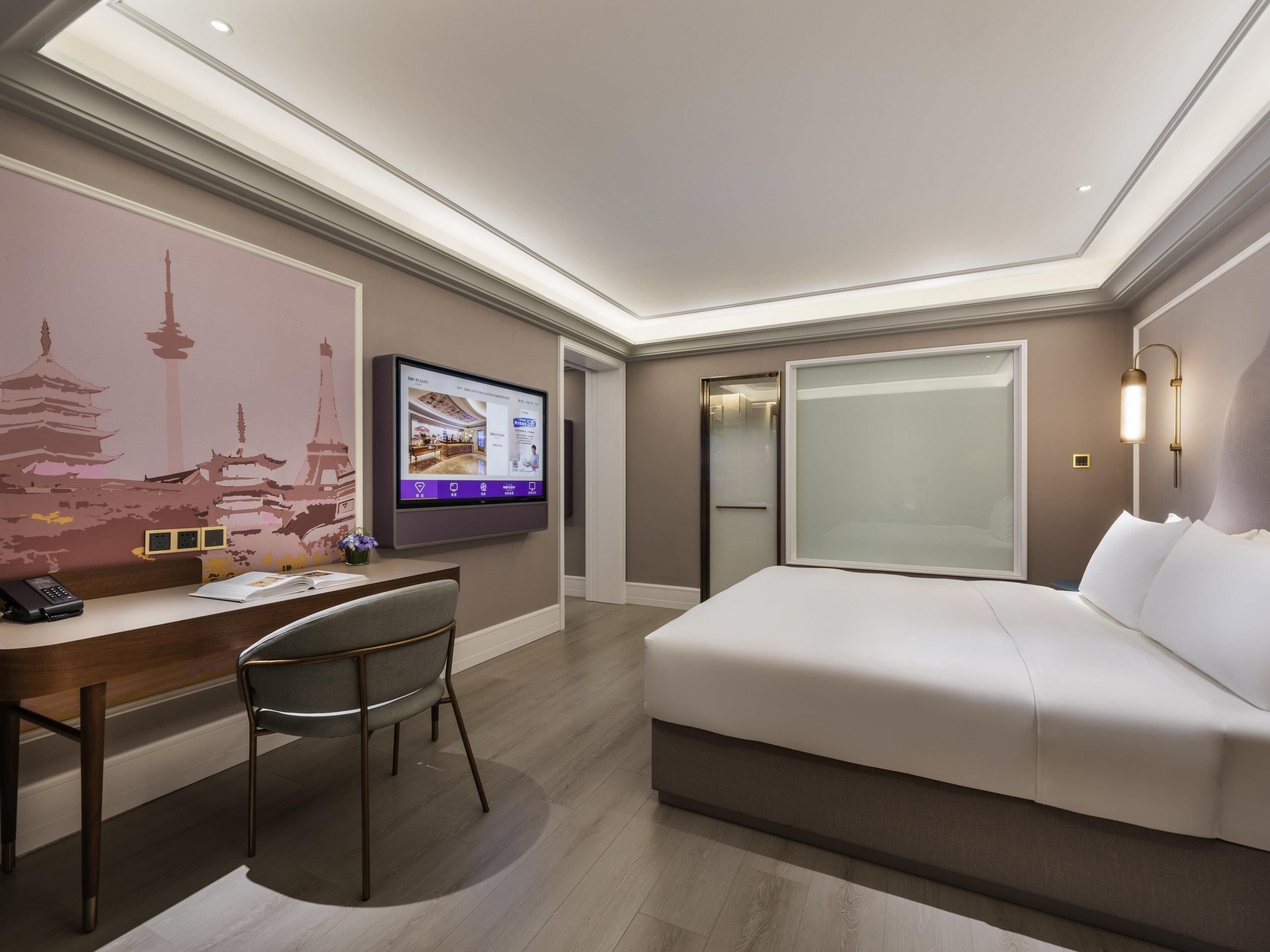 A modern hotel room with a bed, desk, chair, and a TV displaying a room layout.