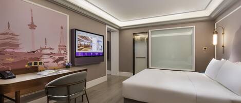 A modern hotel room with a bed, desk, chair, and a TV displaying a room layout.