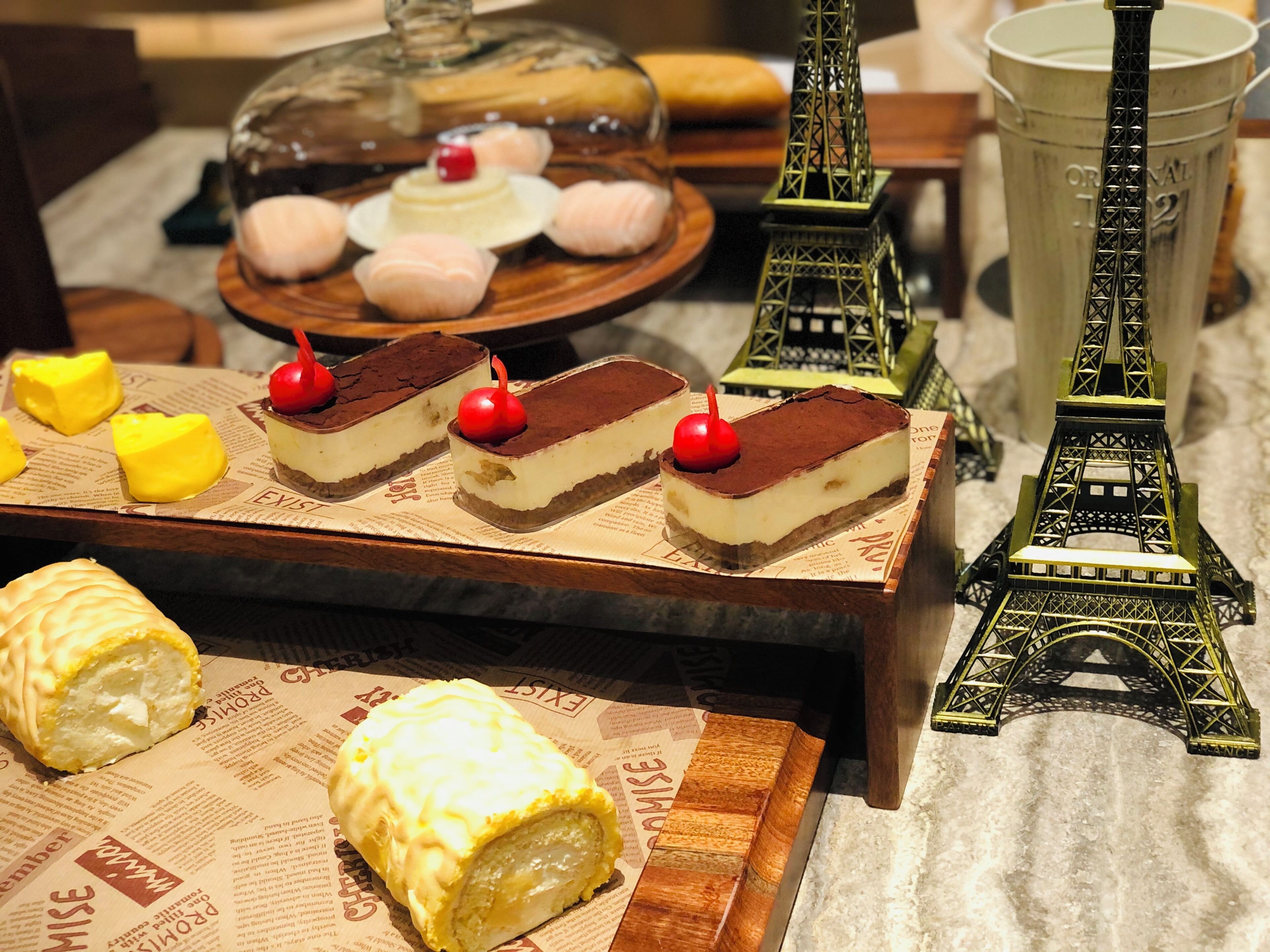 Eine Dessertauslage mit einer Miniatur-Eiffelturms, einer Glaskuppel und einer Auswahl an Desserts vor einer Tageszeitungsoptik.
