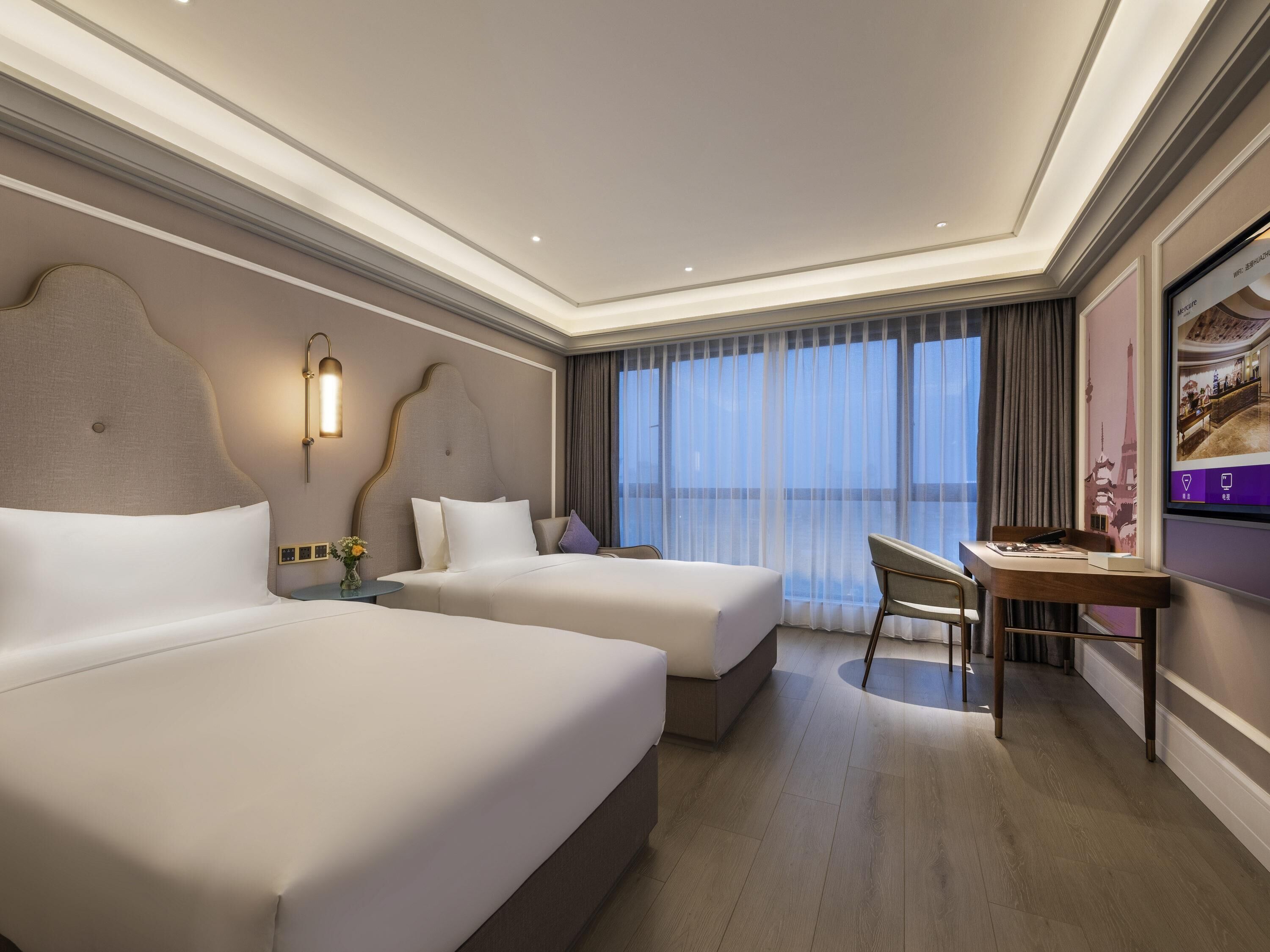 A hotel room with two beds, a desk, a chair, a TV, and a large window with curtains.