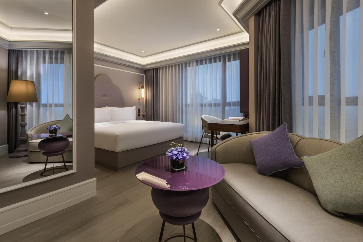 A modern hotel room with a bed, sofa, desk, and a view of the city through large windows.
