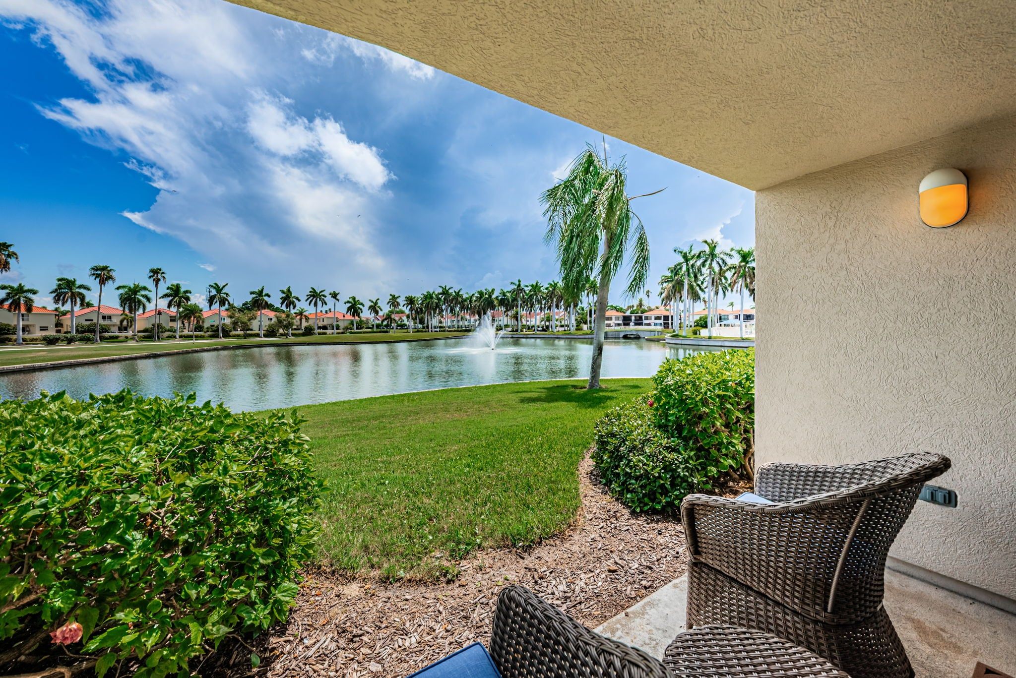 A patio with wicker furniture, a pond, and a view of a resort with palm trees and buildings.