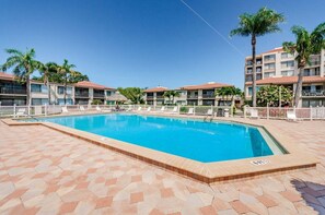 Ein Swimmingpool, umgeben von einer gefliesten Terrasse, mit einem Gebäude im Hintergrund, das Balkone und Palmen zeigt.