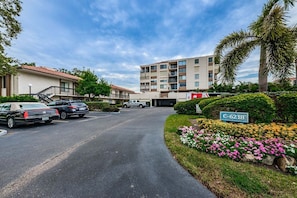 A multi-story residential building with a parking lot, a street sign, and a variety of flowering plants.