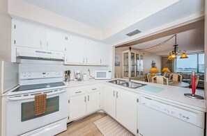 A modern kitchen with white cabinetry, a built-in oven, microwave, and dishwasher. There is a dining area with a view of the city.