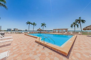 A swimming pool with a tiled deck, lounge chairs, palm trees, and a view of the ocean.