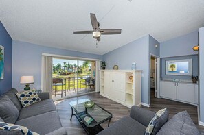 A living room with a ceiling fan, a glass coffee table, grey sofas, and a view of a garden through a sliding glass door.
