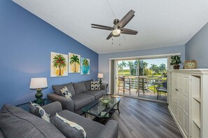 A living room with a blue wall, a ceiling fan, a sofa, a coffee table, and a view of a pool through a sliding glass door. - VVW 6-236 - Vista Verde West - Premium (St. Petersburg)