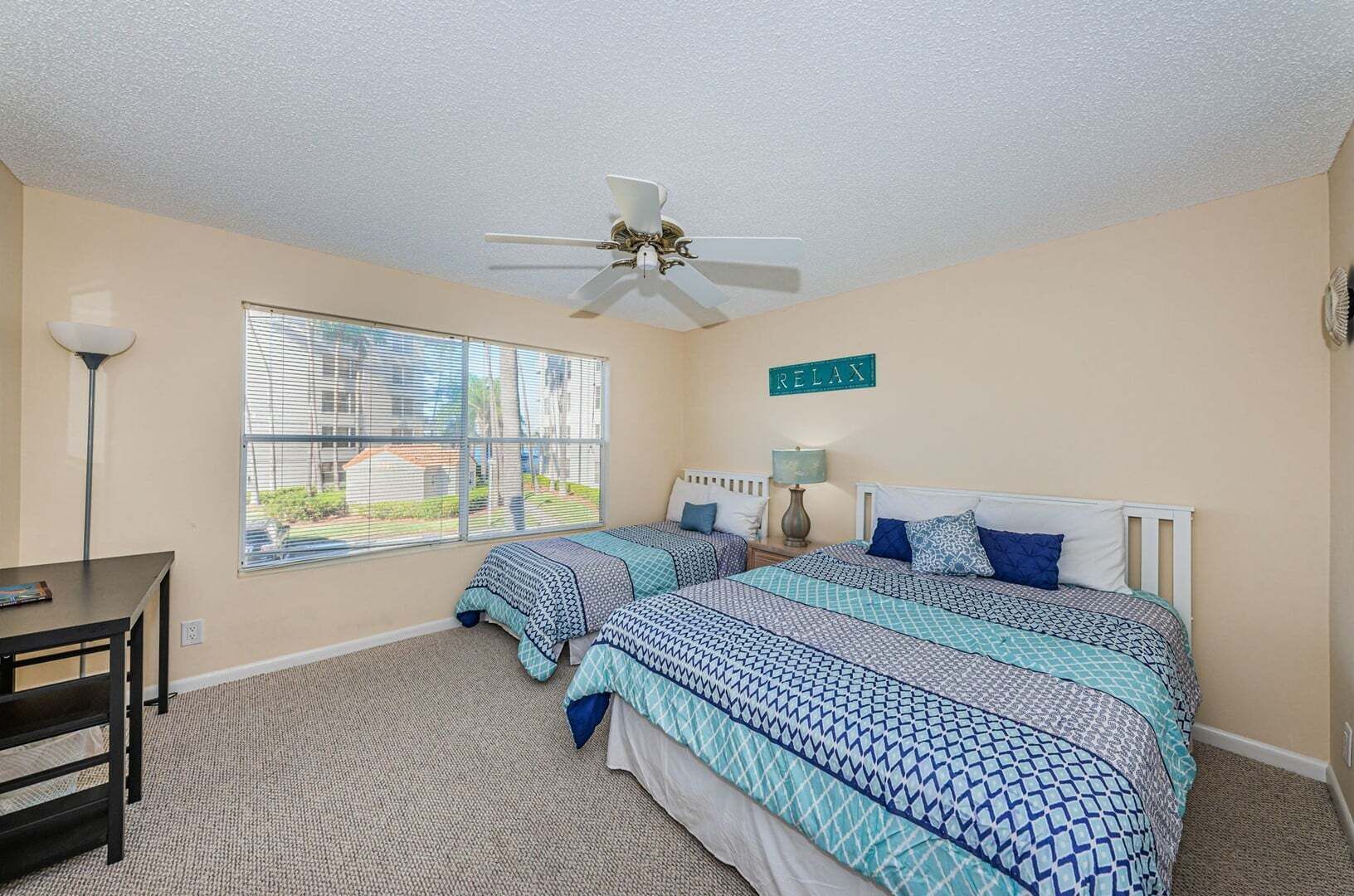 A bedroom with two beds, a ceiling fan, a desk, and a window with blinds.