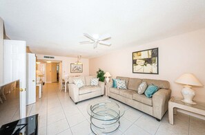 A living room with a ceiling fan, two sofas with patterned cushions, a glass coffee table, and a painting on the wall.