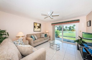 A living room with a sofa, two lamps, a glass coffee table, a ceiling fan, and a view of a golf course through a sliding door.