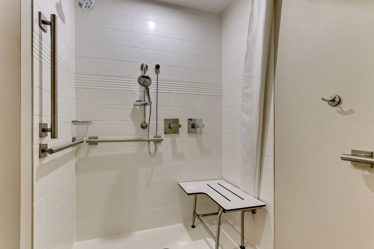 Une salle de bain moderne avec des murs recouverts de carreaux blancs, une douche avec un pommeau de douche amovible et un petit banc blanc.