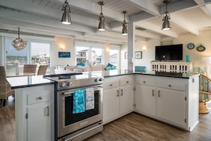 A kitchen with a stove, oven, and white cabinets. There are hanging lights, a television, and a view of the beach.