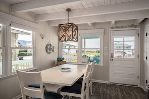 A dining area with a white table and chairs, a hanging light fixture, and a view of the beach through large windows.