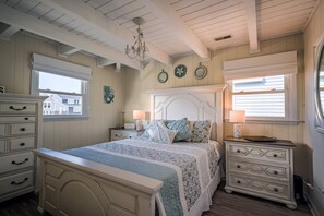 A bedroom with a bed, two bedside tables, a dresser, a chandelier, and a window with blinds.