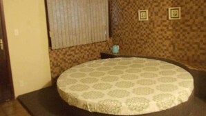 Ein Zimmer mit einem runden Bett, gemustertem Wandpapier, einem Fenster mit Jalousien und einem kleinen Tisch mit einem blauen Gegenstand.