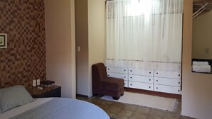 Ein Schlafzimmer mit einem Bett, einem Nachttisch, einem Sessel, einem Fenster mit leichtem Vorhang und einer weißen Kommode.