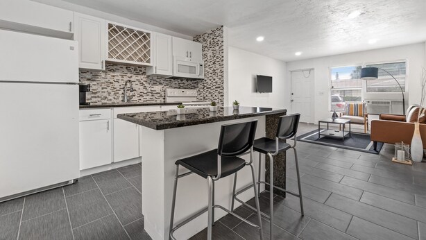 A modern kitchen with white cabinets, a granite countertop, and a built-in microwave. There are two black bar stools and a small dining table with chairs. - Luxury Beach Condo Getaway with fast Wifi (Lake Worth)