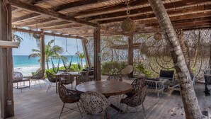 A covered outdoor seating area with a wooden table and wicker chairs, overlooking a beach with palm trees and the ocean.