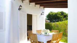 Une terrasse extérieure avec une table blanche et des chaises en bois, entourée de murs blancs et d’une pergola en bois.