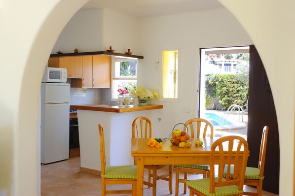 Cocina compacta con microondas, refrigerador y armarios de madera. Una mesa de comedor con sillas está ubicada cerca de una ventana con vista a una piscina.