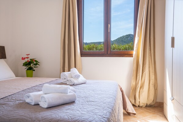 A neatly made bed with rolled towels, a green potted plant, and a window with curtains.