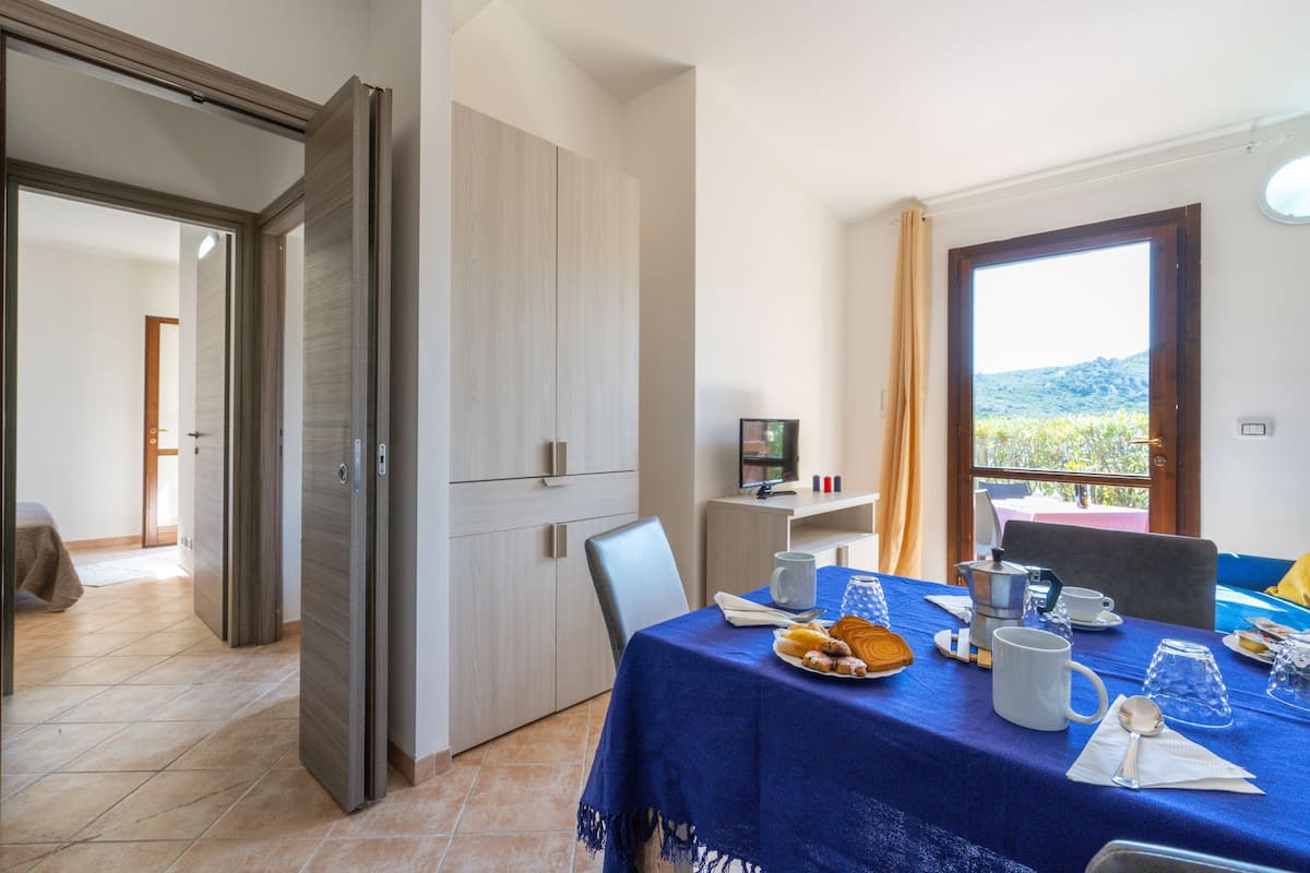 A dining area with a table set for two, a view of the outdoors, and a kitchen area visible in the background.