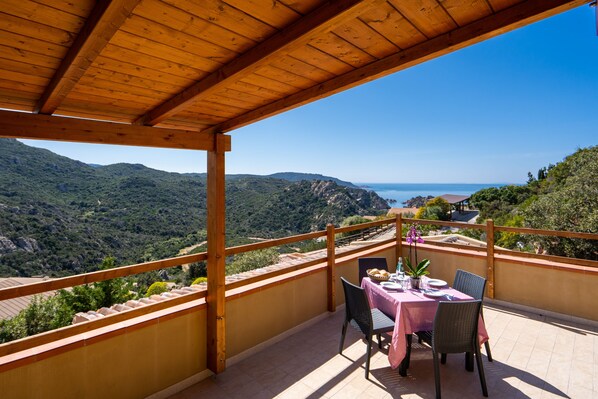 A wooden balcony with a table set for two, overlooking a mountainous landscape and the sea.