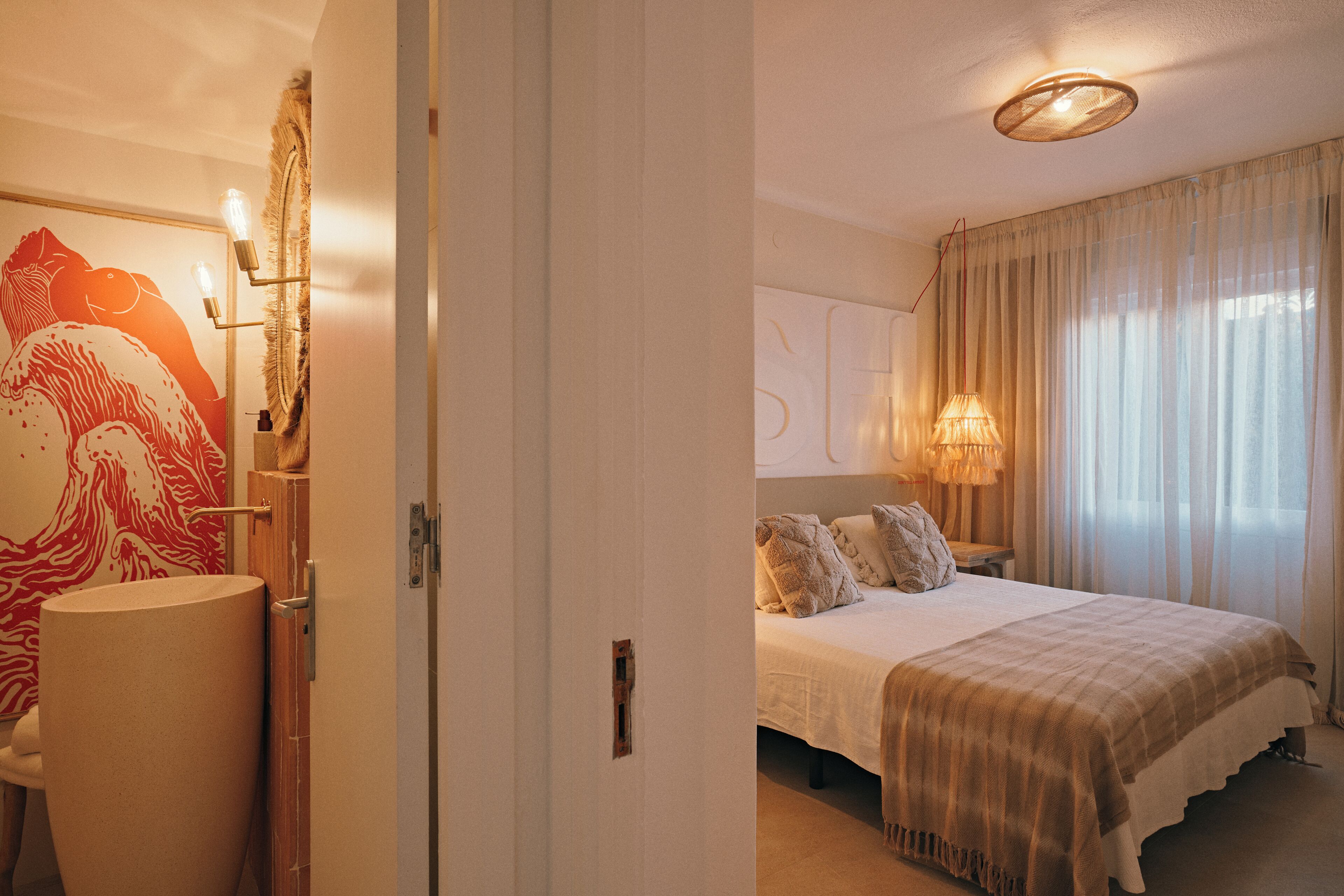 Quarto de hotel com uma cama grande, uma mesinha de cabeceira, um abajur e uma parede com uma pintura abstrata vermelha.