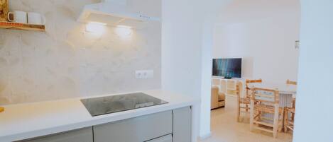 A modern kitchen with a white countertop, grey cabinets, and a built-in stove. There is a wooden shelf with cups and a television in the background.