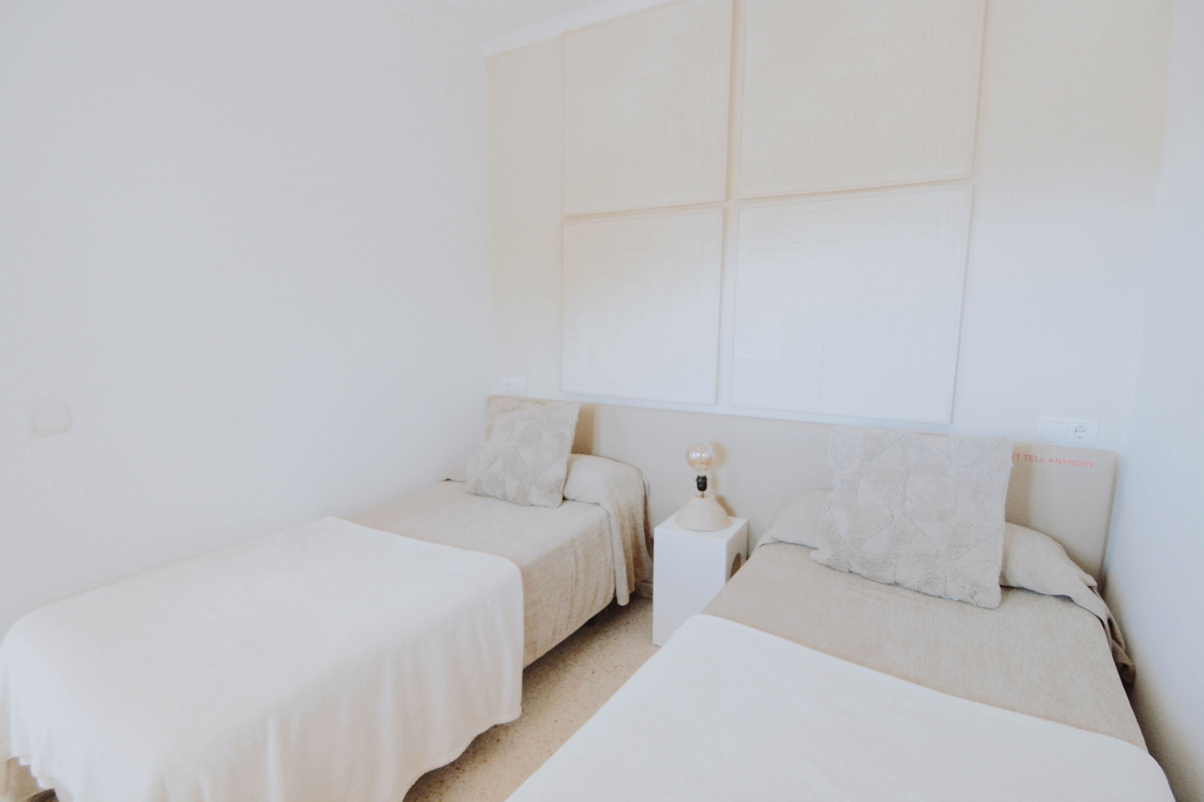 A minimalist bedroom with two single beds, a nightstand with a lamp, and a plain white wall.