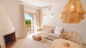 A living room with a sofa, a round table, and a fireplace. There is a balcony with curtains and a view of greenery. - Secret Oasis Ibiza- Adults Only (Sant Josep de sa Talaia)