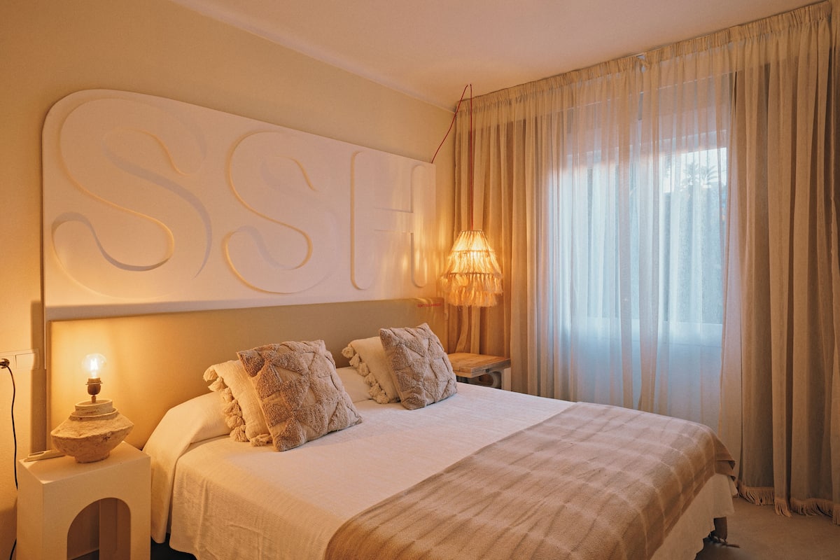 Una cama bien hecha con una gran obra de arte iluminada sobre ella, una lámpara de noche y vistas a una ventana con cortinas translúcidas.