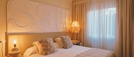 Una cama bien hecha con una gran obra de arte iluminada sobre ella, una lámpara de noche y vistas a una ventana con cortinas translúcidas.