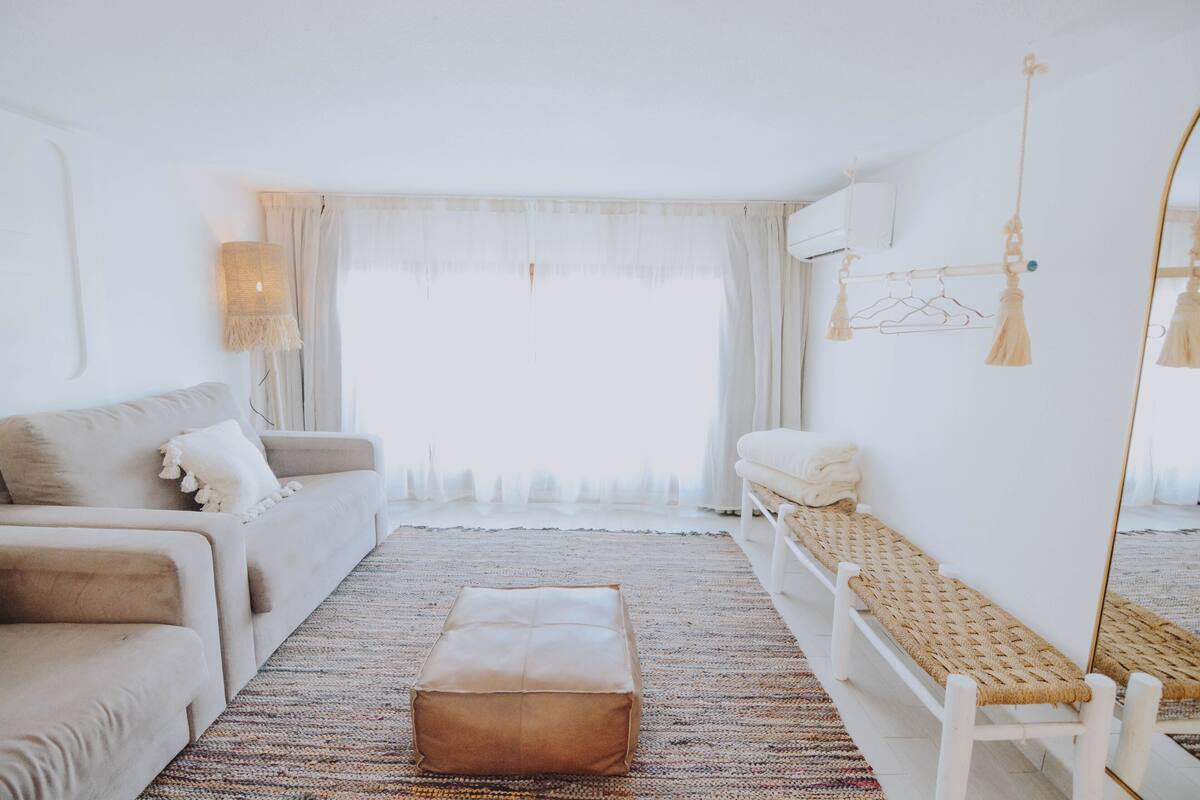 Una sala moderna con sofá, reposapiés, banco y un amplio ventanal con cortinas.