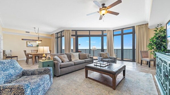 A spacious living room with a large window, a ceiling fan, and a view of the ocean. - New to the Rental Market - Lazy River and Slide! (Orange Beach)