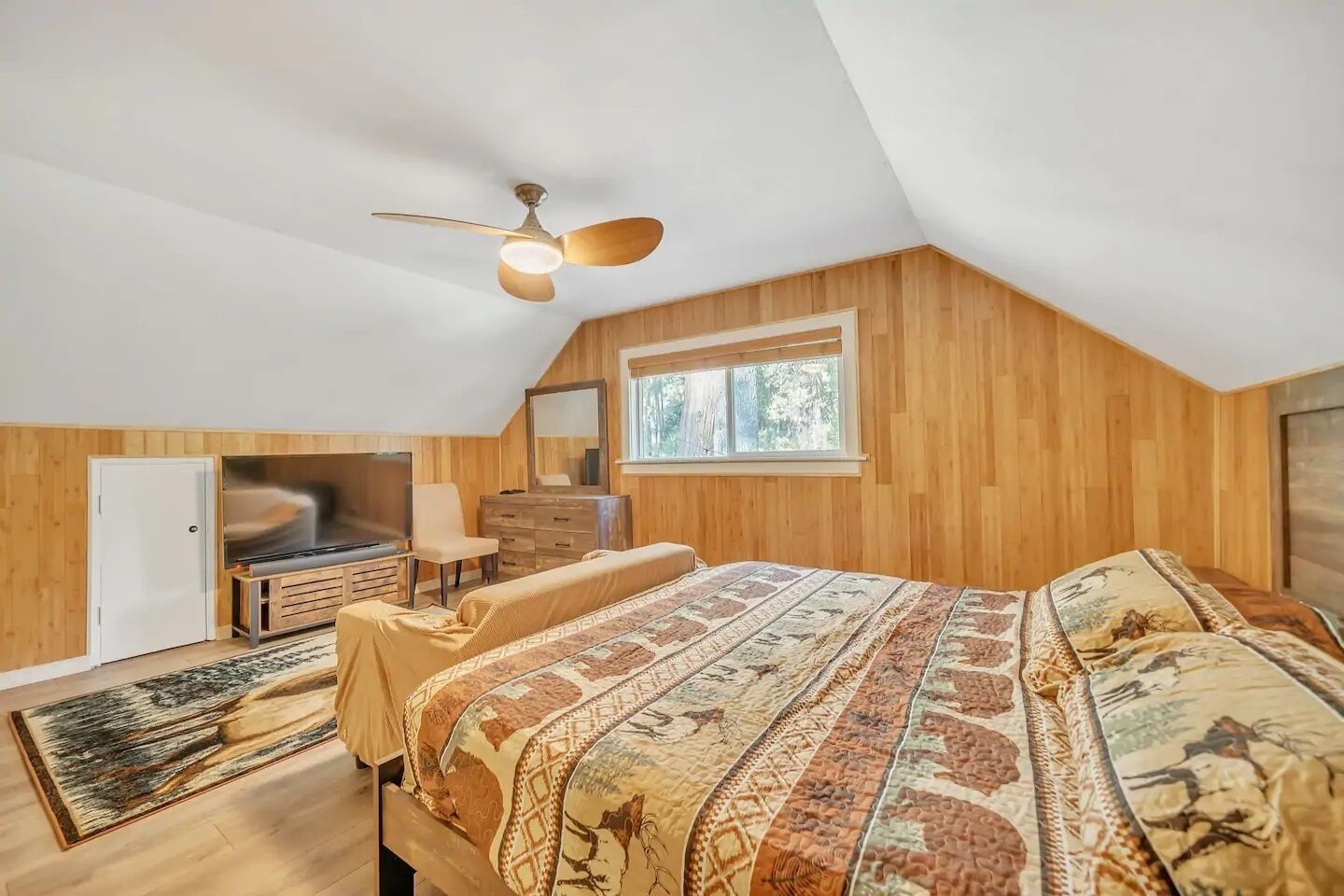 Une chambre avec un plafond en pente, un lambris en bois, un lit avec une literie à motifs, une commode, une chaise, un téléviseur et une fenêtre.