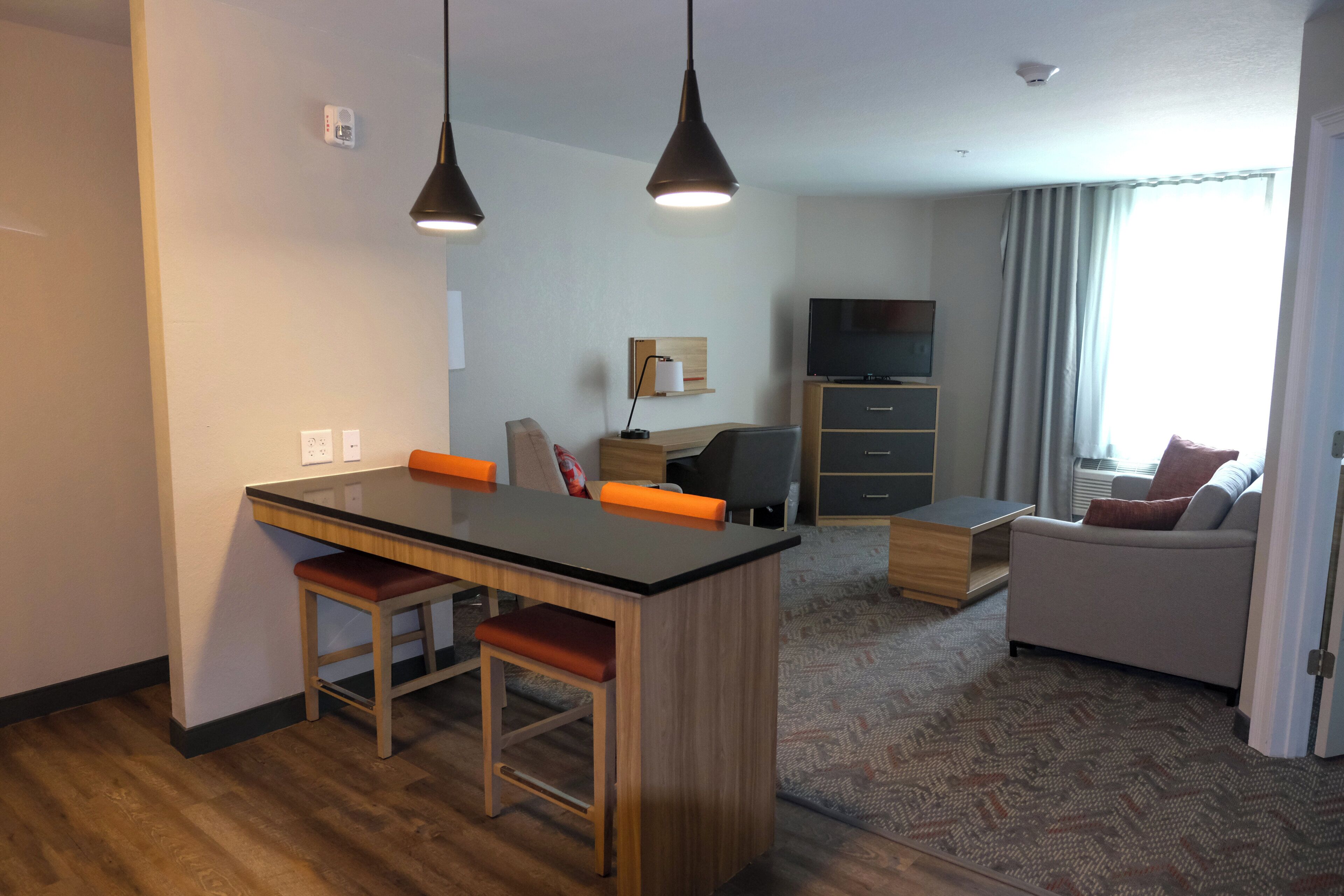A modern hotel room with a kitchenette, a dining area, a sofa, and a TV.