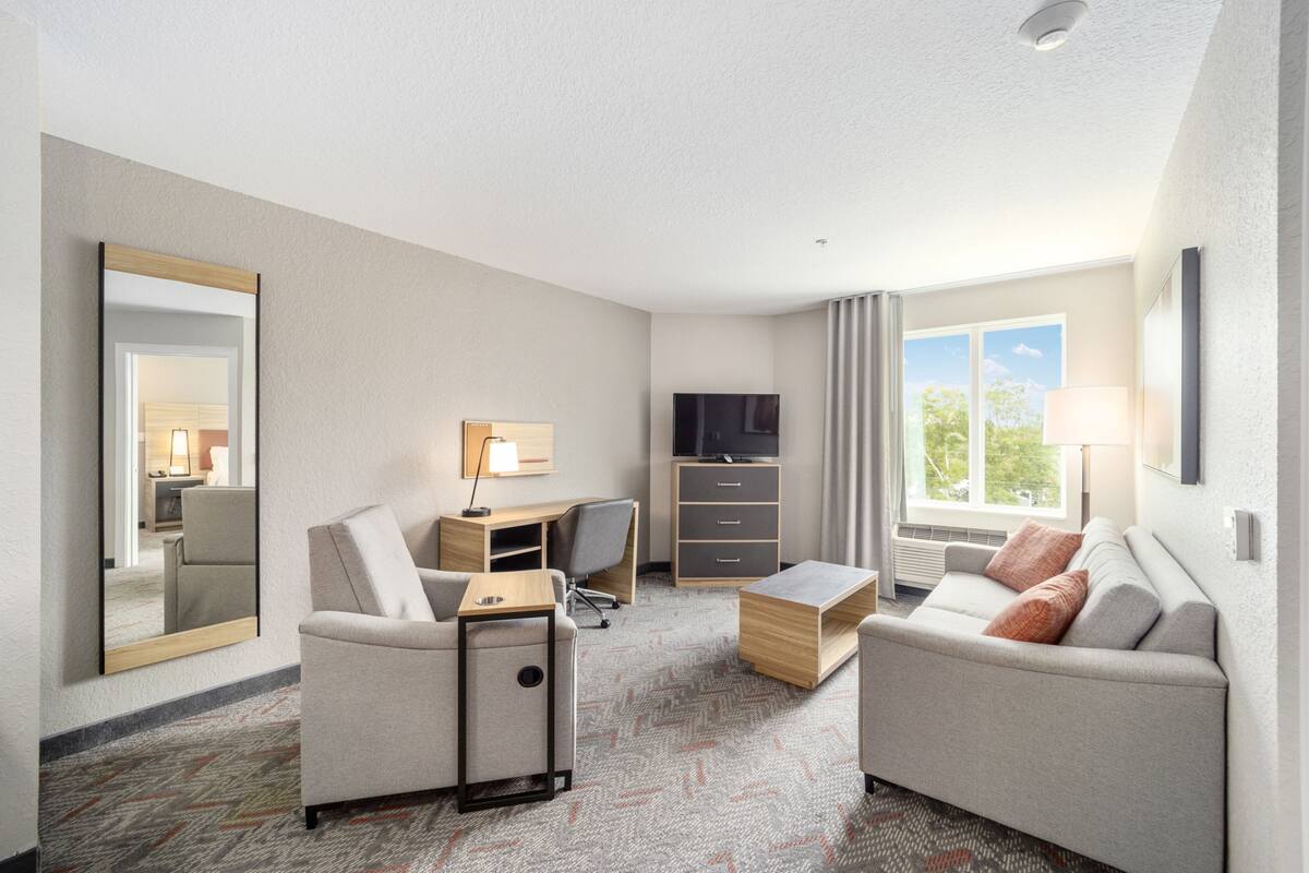 Habitación de hotel con sofá, escritorio y silla, televisión, espejo y ventana con cortinas.