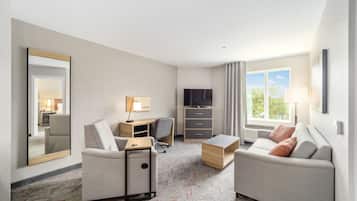 A hotel room with a sofa, a desk with a chair, a TV, a mirror, and a window with curtains.