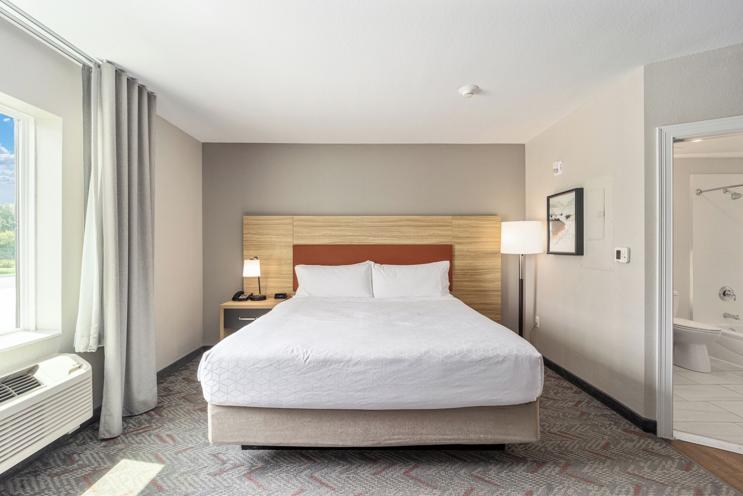 A hotel room with a large bed, a bedside table with a lamp, a window with curtains, and a bathroom visible through an open door.