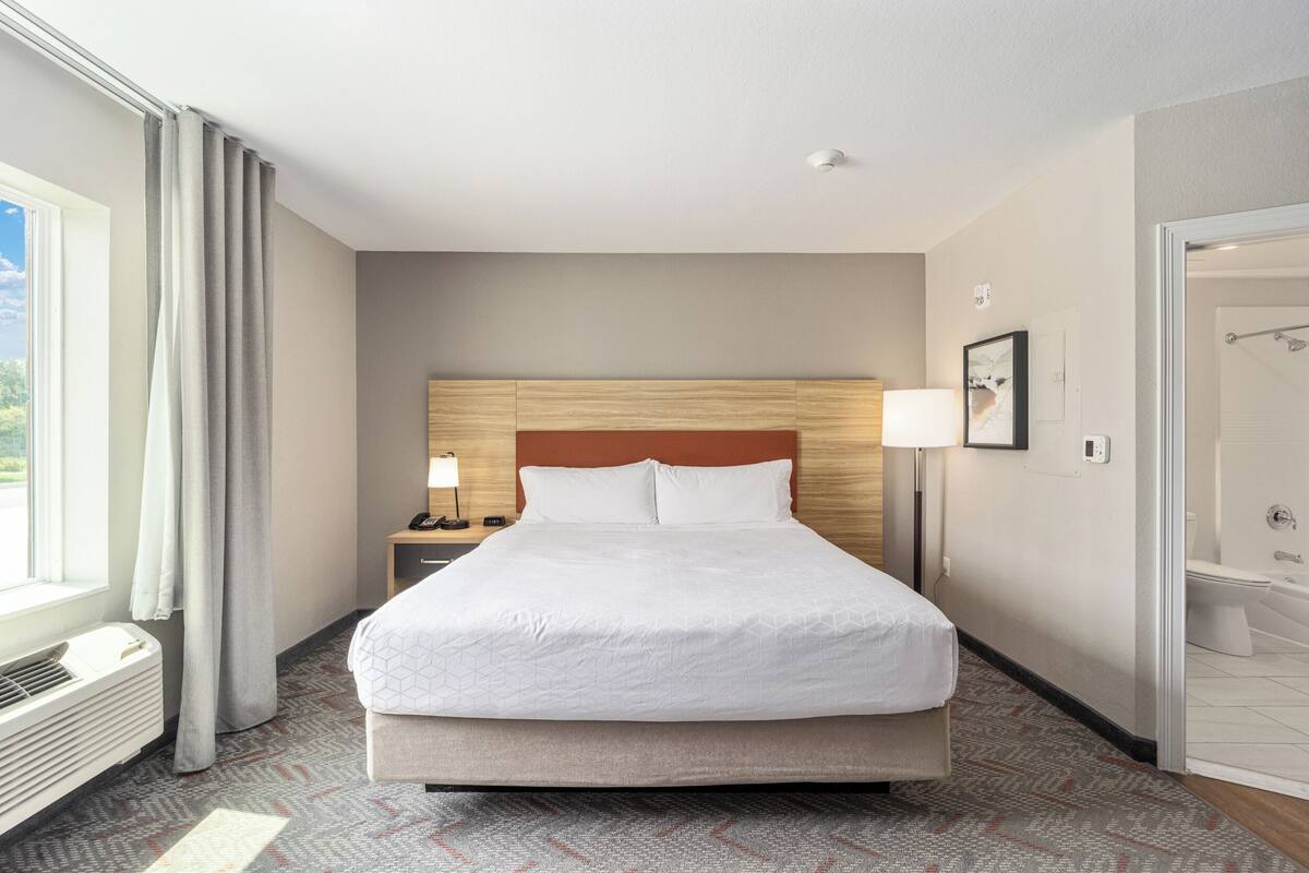 Ein Hotelzimmer mit einem großen Bett, einem Nachttisch mit Lampe, einem Fenster mit Vorhängen und einem durchgegangene Tür erkennbaren Badezimmer.