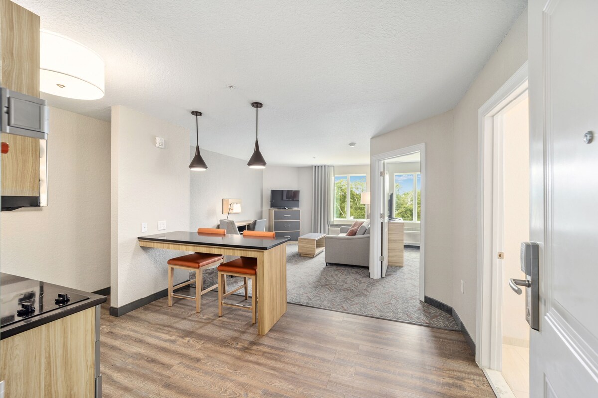 A modern kitchen with a central island, pendant lighting, and a view to a living area with a sofa and a window.